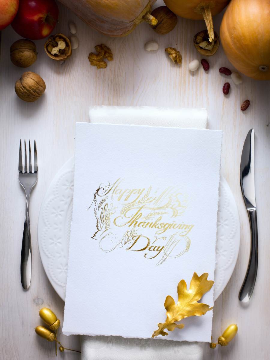 image presents a refined Thanksgiving table setting centered around a napkin elegantly embossed with golden Happy Thanksgiving Day lettering accompanied by a subtle hint of autumn with golden-painted acorns and a few walnuts suggesting a sophisticated yet festive atmosphere