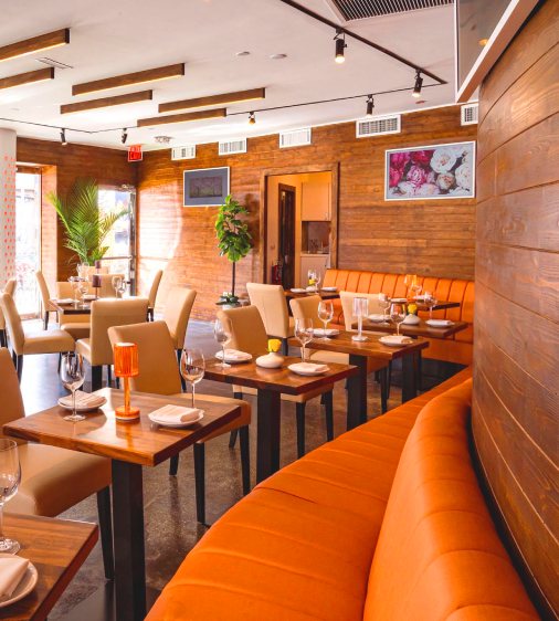 A long orange booth lines the wall paired with wooden tables that are neatly set for dining. Each table is accompanied by cream colored restaurant chairs